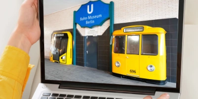 Eine Person hält einen Laptop, auf dessen Bildschirm der Eingang des U-Bahn-Museums Berlin mit blauem Schild und zwei gelben historischen U-Bahn-Wagenfronten zu sehen ist.