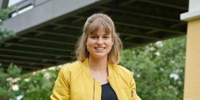 Auszubildene Michèle steht draußen vor einer großen Brücke und lächelt in die Kamera.