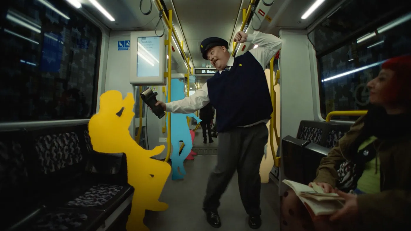 Ein Filmausschnitt ist zu sehen, der einen Kontrolleur in einer U-Bahn zeigt. Er scheint zu tanzen während er die Passagiere kontrolliert, die teils bereits nur als farbige Silhouetten zu erkennen sind.