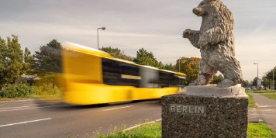 Im Vordergrund ist eine Statue des Berliner Bären zu sehen, im Hintergrund ein fahrender Bus der BVG.