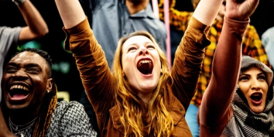 Jubelnde Menschen die die Arme in die Luft strecken, der Fokus liegt auf einer jungen blonden Frau.