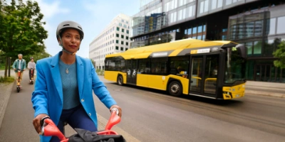 Eine Frau in einem blauen Blazer fährt Fahrrad, auf der gegenüberliegenden Straße fährt ein Bus der BVG vorbei.