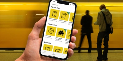 A hand holds a smartphone on which the ticket app can be seen. In the background there is a passing subway train.