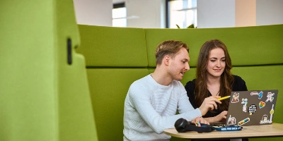 Zwei Personen sitzen an einem Tisch und schauen auf einen Laptop.