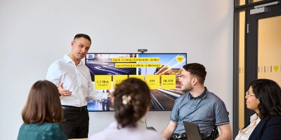 Mehrere Personen sitzen in einem Besprechzungsraum gemeinsam vor einem Bildschirm, auf dem eine Präsentation zu sehen ist.