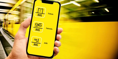 Eine Hand hält ein Smartphone auf dem die verschiedenen Icons der BVG-App-Familie abgebildet sind. Im Hintergrund ist verschwommen eine U-Bahn zu sehen.