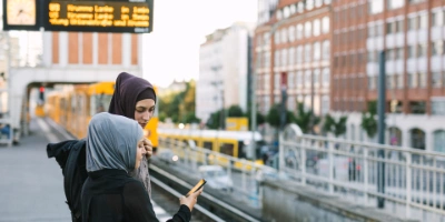 Zwei Mädchen mit Kopftuch stehen an einer Haltestelle und schauen auf ein Handy.