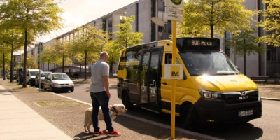 Eine Person mit einem Blindenhund wartet an einer Haltestelle, während gerade ein BVG Muva Kleinbus einfährt.