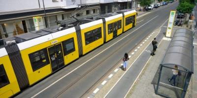 Eine Tram fährt in eine Haltstelle ein. Gegenüber warten Menschen.