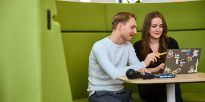 Zwei Personen sitzen auf einer grünen gepolsterten Bank und schauen auf einen Laptop.