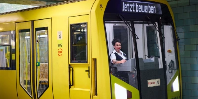 Eine U-Bahnfahrerin sitzt in der U-Bahn und hält am Bahnsteig. Die Anzeige zeigt "Jetzt bewerben" an.