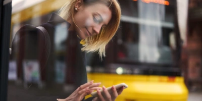 Eine Frau tippt auf ihrem Smartphone, im Hintergrund fährt eine Tram der BVG vorbei.