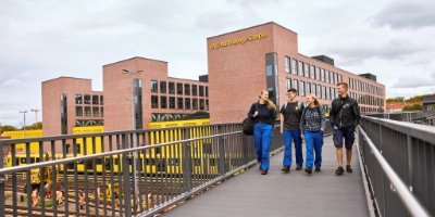 Vier Auszubildende der BVG gehen lachend über eine Fußgängerbrücke mit Blick auf den BVG-Ausbildungs-Campus in Berlin; im Hintergrund sind gelbe Arbeitsfahrzeuge und Gleisanlagen zu sehen.