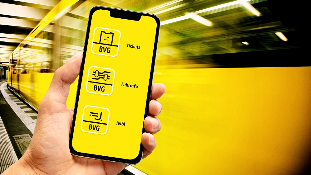 Eine Hand hält ein Smartphone auf dem die verschiedenen Icons der BVG-App-Familie abgebildet sind. Im Hintergrund ist verschwommen eine U-Bahn zu sehen.