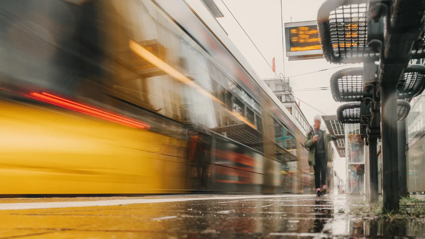 Eine Tram verlässt die Haltstelle. Es regnet. Das in der Froschperspektive aufgenommene Bild der Haltestelle ist geprägt von Bewegungsunschärfe und starken Lichtreflexionen durch den Regen.