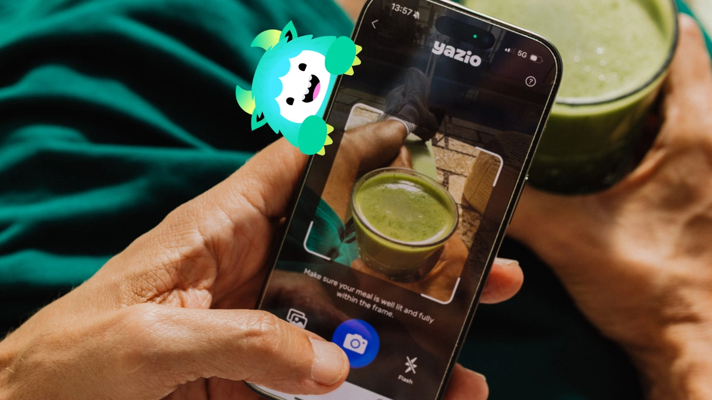 Es ist ein Handxbildschirm zu sehen, über den ein grüner Smoothie fotografiert wird, um ihn in der Yazio-App einzutragen. Am Handy hängt ein kleines grün-weißes Comic-Monster. 