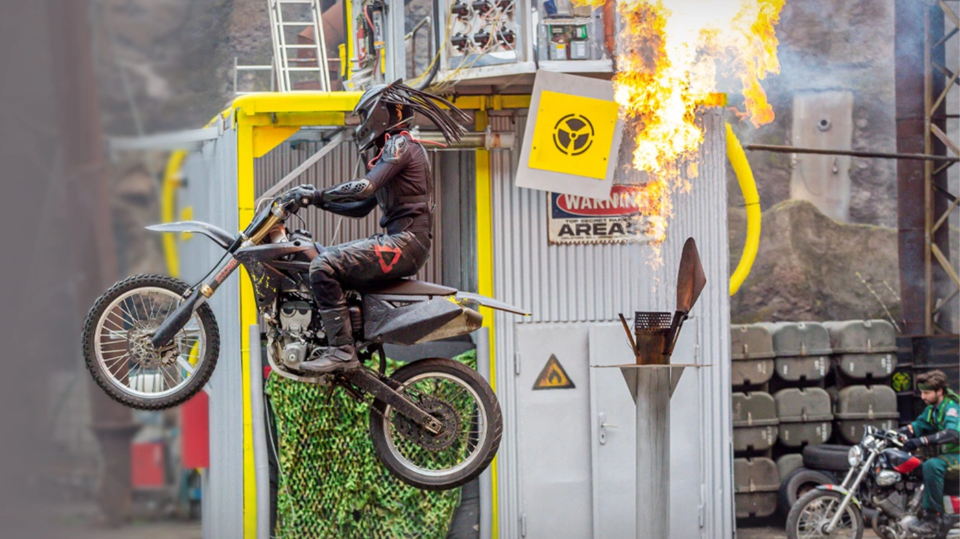 Vor einer Kulisse im Filmpark Babelsberg ist ein Motorradfahrer zu sehen, der durch die Luft fliegt - im Hintergrund schlagen Flammen hoch.