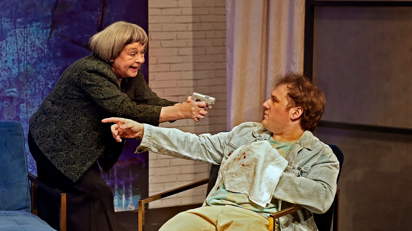Ein Szenenbild aus einem Theaterstück: Eine ältere Frau richtet eine Waffe auf einen Mann, der auf einem Stuhl sitzt.