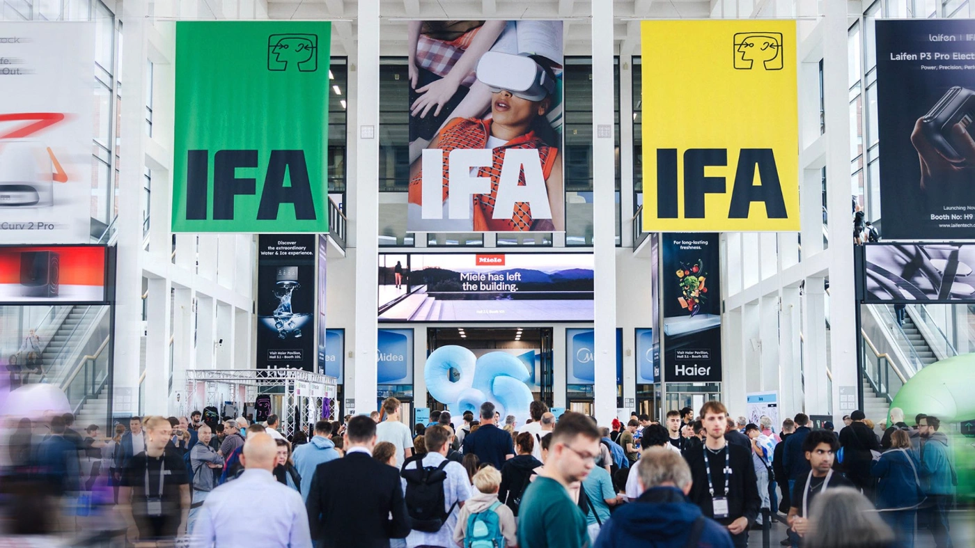 Eine belebte Messehalle der IFA mit großen farbigen Bannern und zahlreichen Besucherinnen und Besuchern. Im Vordergrund bewegen sich Menschen durch den Eingangsbereich, während im Hintergrund Ausstellungsstände und Werbeflächen für Technikunternehmen zu sehen sind.