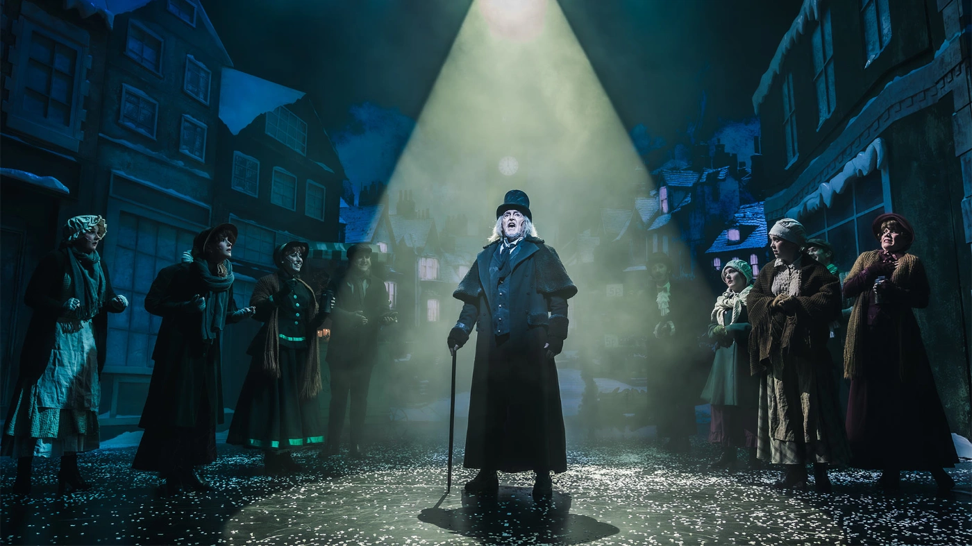 Ein Schauspieler in schwarzer Kleidung auf einer Bühne im Rampenlicht. Um ihn herum stehen weitere Schauspieler in einer dunklen winterlichen Atmosphäre.