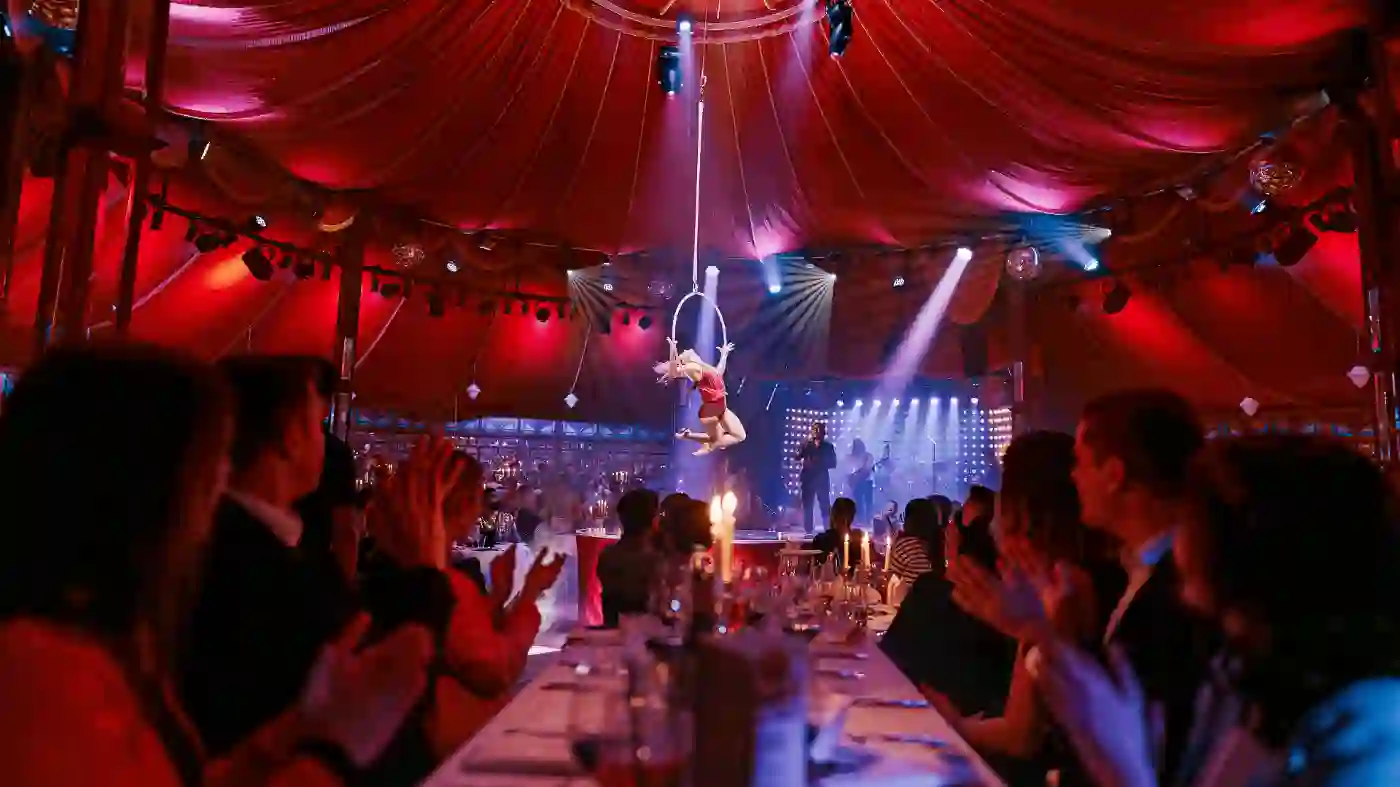 Der Blick von einem aus in die Mitte eines Zirkuszelts, wo eine Akrobatin an einem Ring in der Luft turnt.