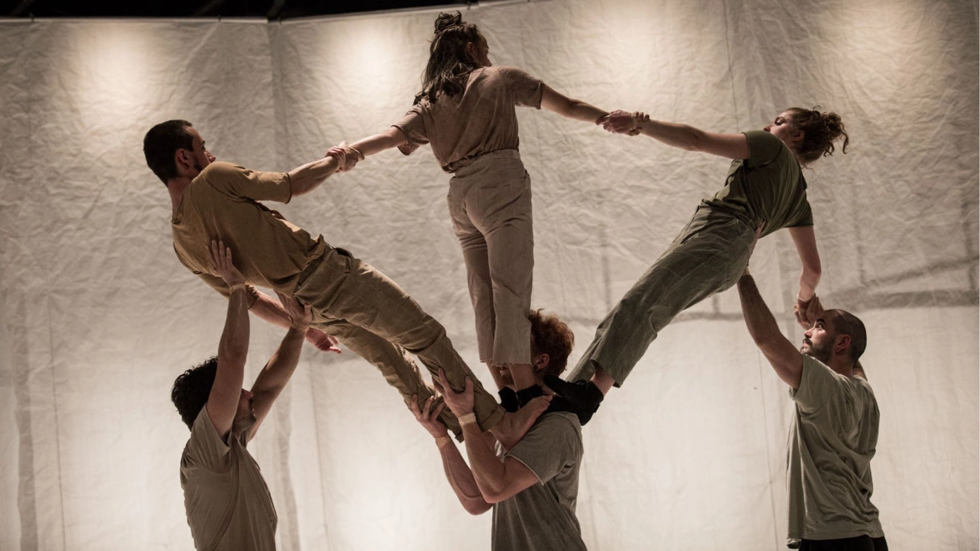 6 Aktobat*innen in beigen Outfits si8nd zu sehen; dabei stehen drei Personen auf der Schulter einer anderen Person, werden gestützt und bilden dabei einen Halbkreis. 