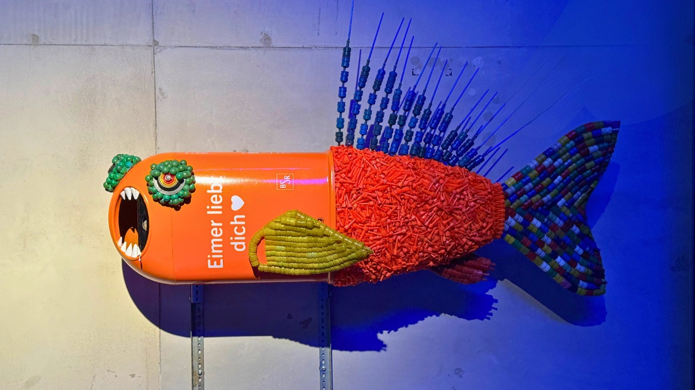 Aus einem orangenen Mülleimer wurde eine Skulptur gebaut, die wie ein Fisch mit aufgerissenem Maul aussieht: Dazu wurde Müll und Plastikteile benutzt. 