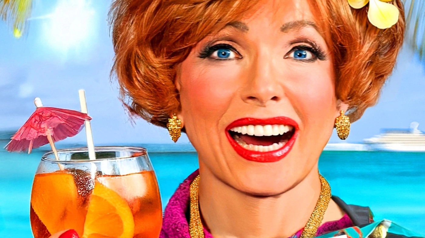 Lachende Frau mit rotem Haar und rotem Lippenstift hält einen Cocktail vor einer tropischen Meereskulisse.