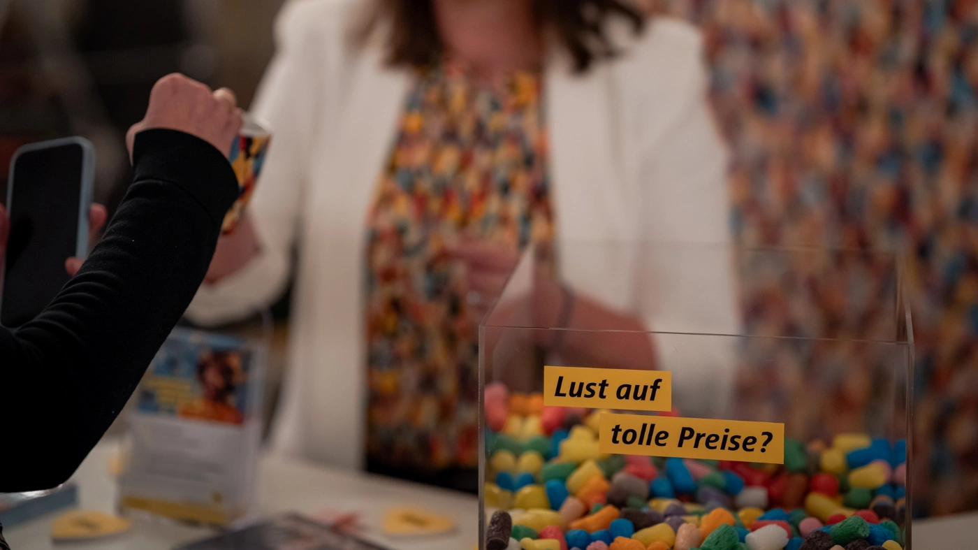 Nahaufnahme eines Tresens mit einer transparenten Box voller bunter Süßigkeiten und dem Text „Lust auf tolle Preise?“. Im Hintergrund stehen unscharf Personen an einem Infostand.