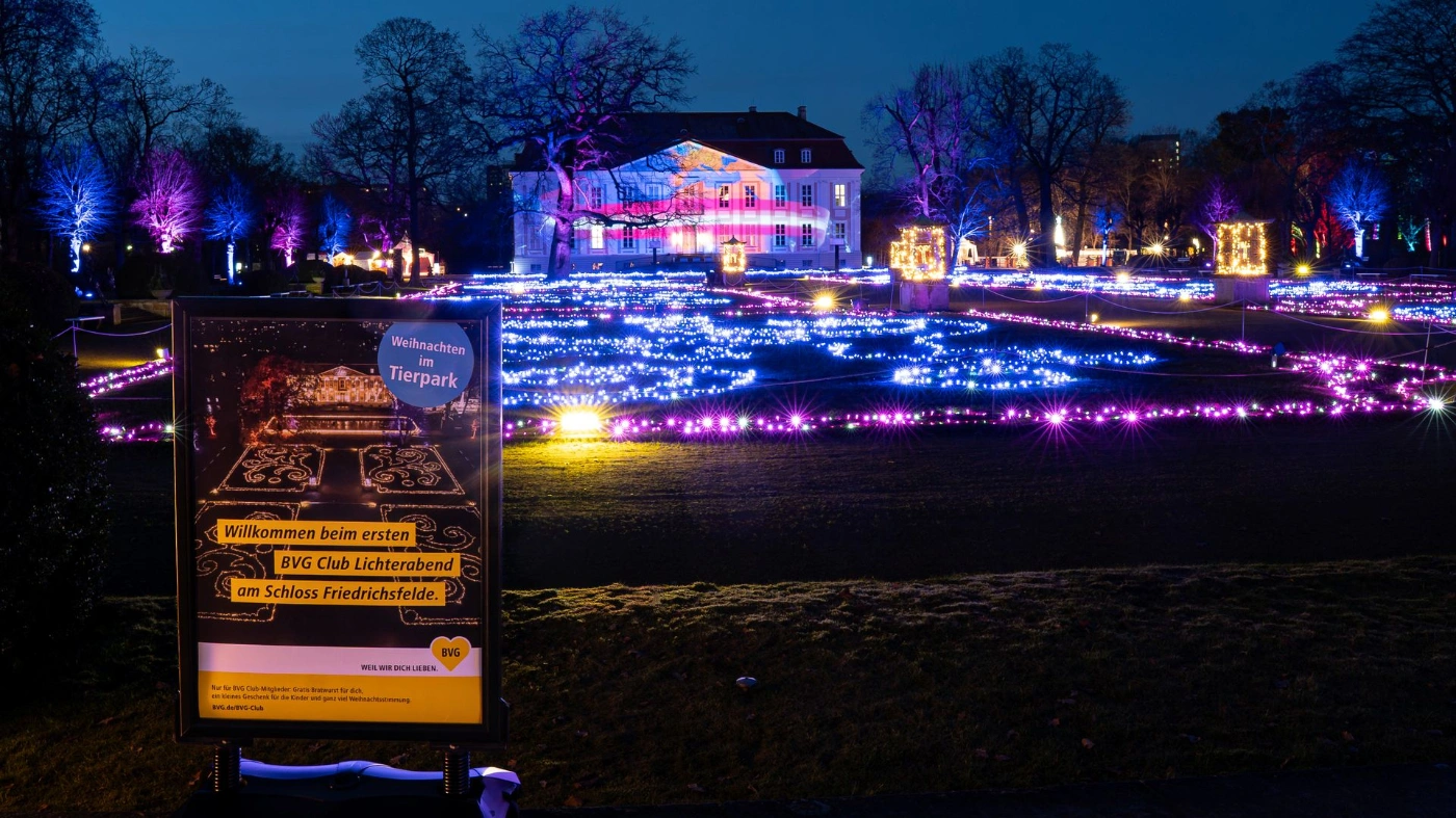 Nächtlicher Park mit bunten Lichterketten und illuminiertem Schloss im Hintergrund. Im Vordergrund steht ein Schild zum „Weihnachten im Tierpark“ und BVG Club Lichterabend.