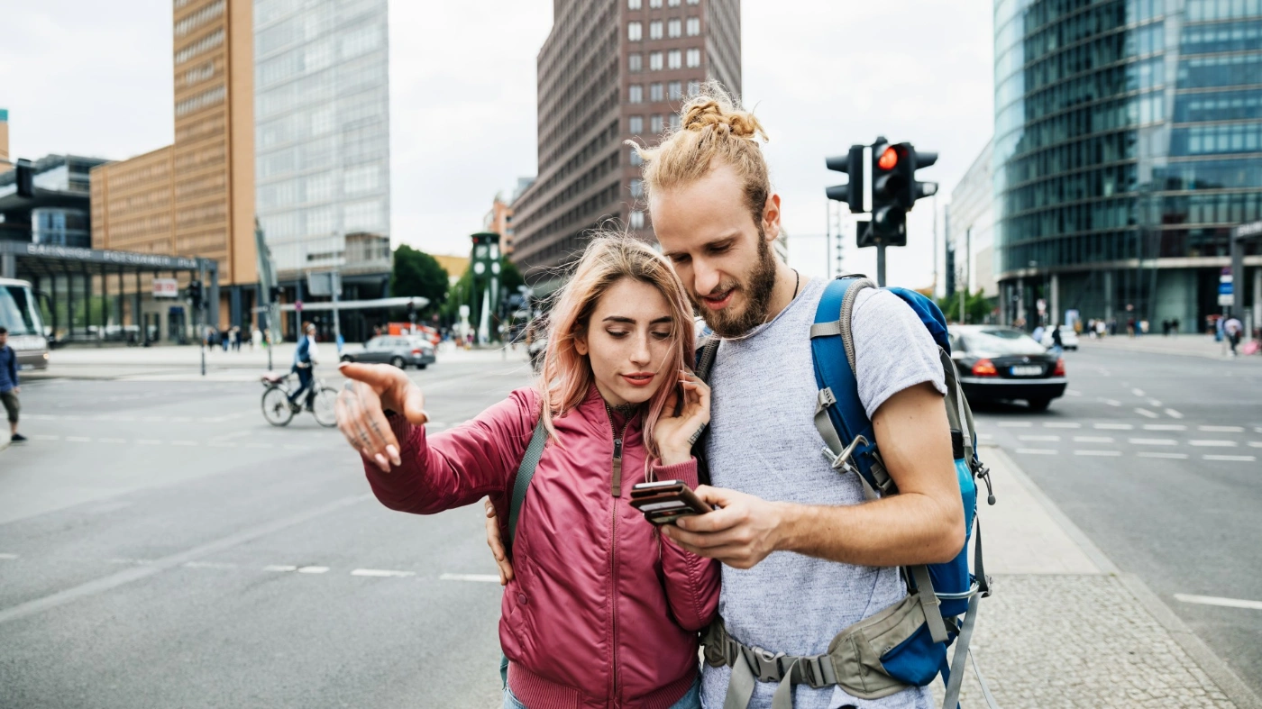 zwei Personen sind an der Kreuzung einer Großststadt zu sehen: Eine junge Frau und ein junger Mann, sie schauen gemeinsam auf ein Smartphone. Die Frau zeigt mit dem Finger n die Richtung, in die sie voraussichtlich müssen.