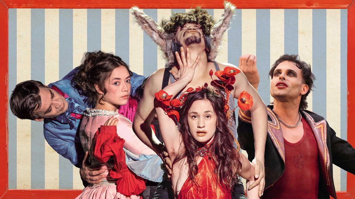 Vier Personen in fantasievollen Theaterkostümen posieren vor einem gestreiften Hintergrund. Eine Figur trägt auffällige Tierohren, andere sind mit Blumen und dramatischem Make-up geschmückt.