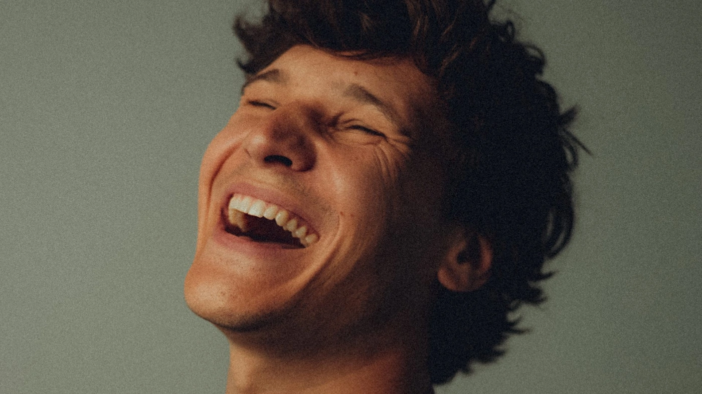 Nahaufnahme eines lachenden jungen Mannes mit geschlossenen Augen und dunklen Haaren vor neutralem Hintergrund. Es handelt sich bei dem Mann um Wincent Weiss.