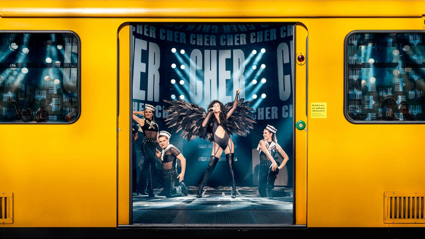 Es geöffnete gelbe U-Bahn zeigt einen Ausschnitt der Cher-Show, wobei eine Künstlerin im Scheinwerferlicht die Sängerin Cher verkörpert. Sie trägt ein schwarzes Bühnenkostüm mit schwarzen Flügeln, um sie herum stehen Tänzer*innen. 