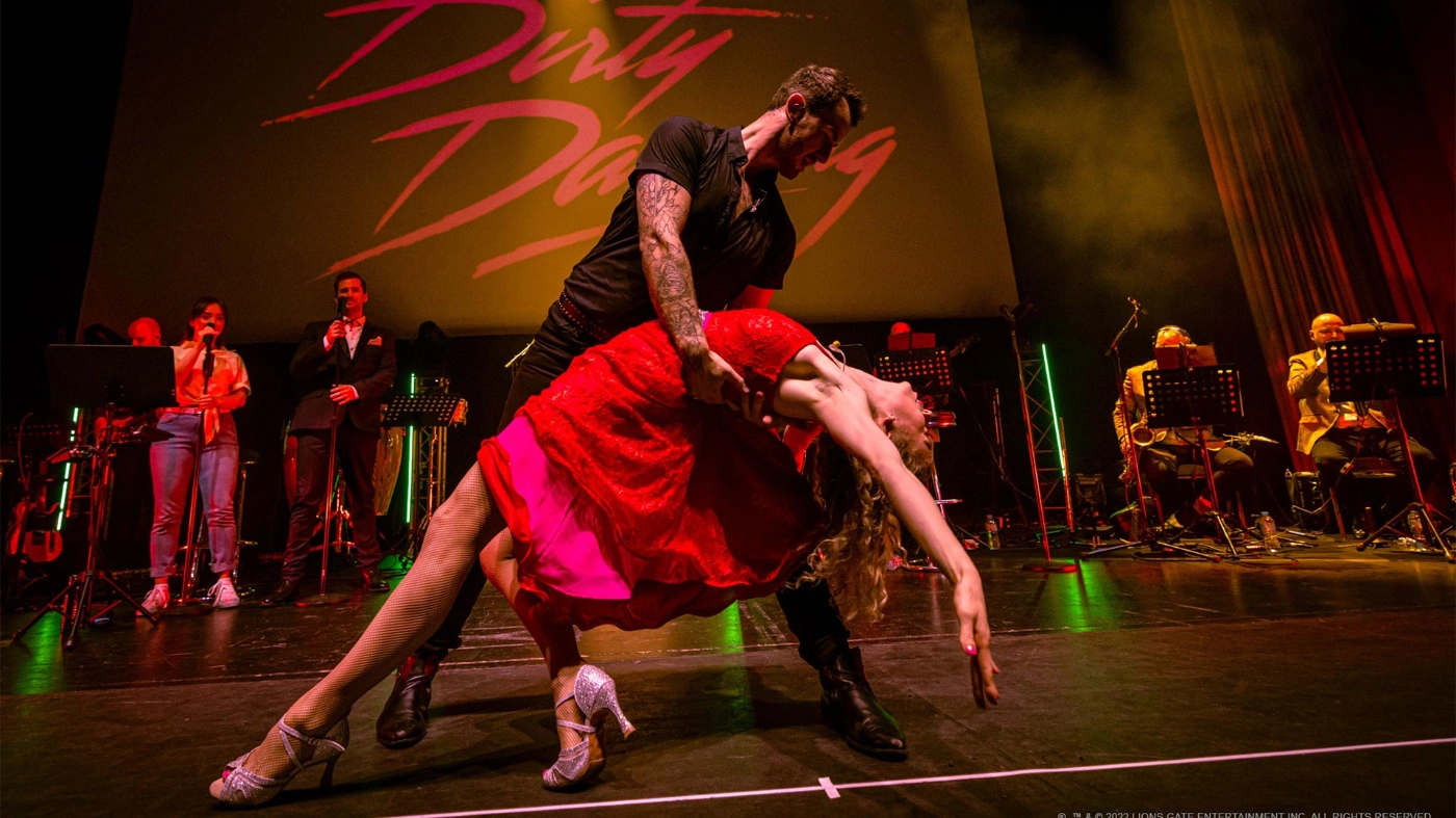 Zwei Tänzer sind zu sehen: Der männliche Tänzer, schwarz gekleidet, hält eine weibliche Tänzerin, in einem roten Kleid vor Musiker*innen.