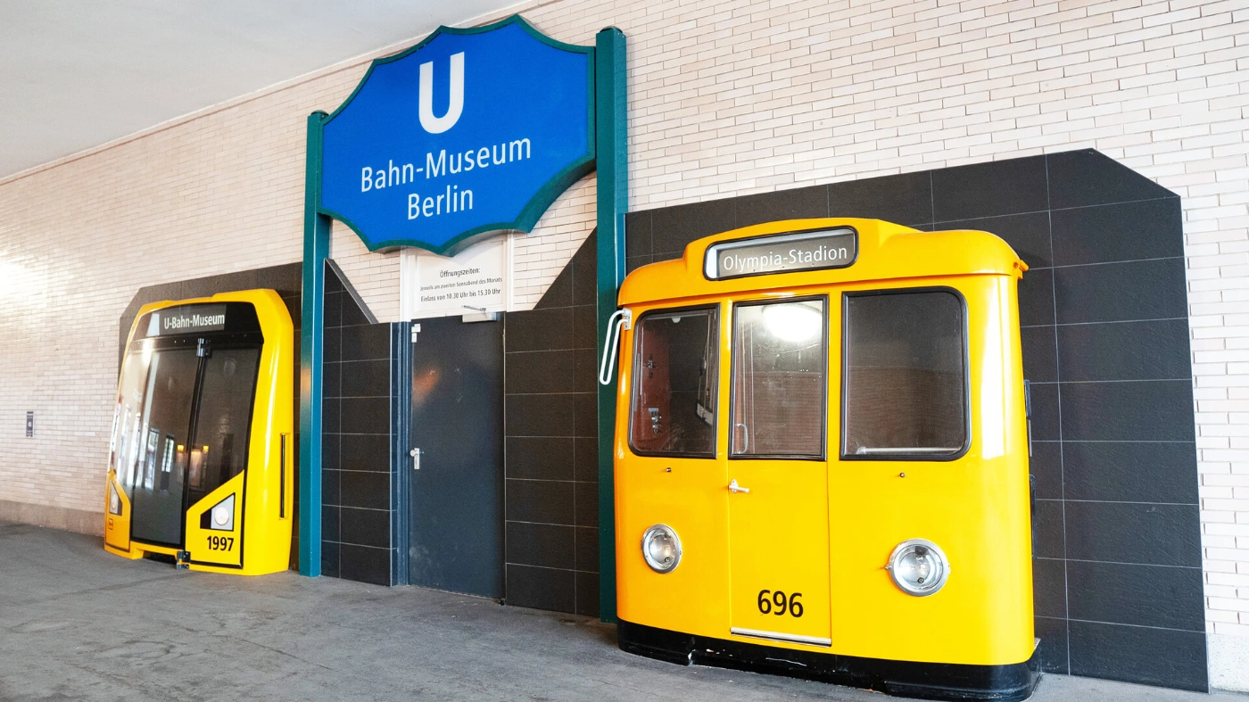 Eingang zum U-Bahn-Museum Berlin mit großem blauem Schild an einer hellen Backsteinwand. Daneben stehen zwei gelbe Frontteile historischer U-Bahn-Wagen, eines mit Zielanzeige „Olympia-Stadion“.