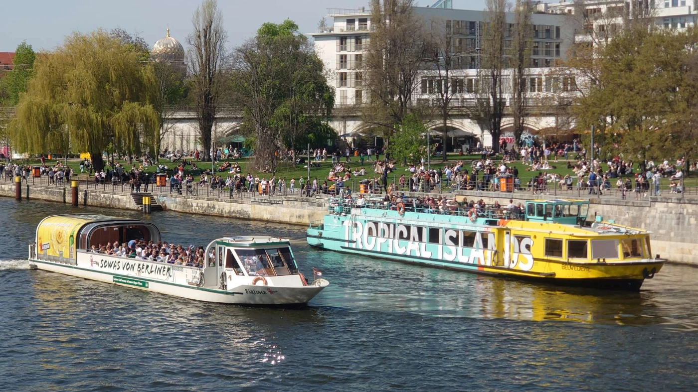 Zwei Ausflugsschiffe fahren auf der Spree an einem belebten Ufer mit vielen Menschen auf Wiesen und Promenade vorbei. Im Hintergrund stehen Bäume und Wohngebäude.