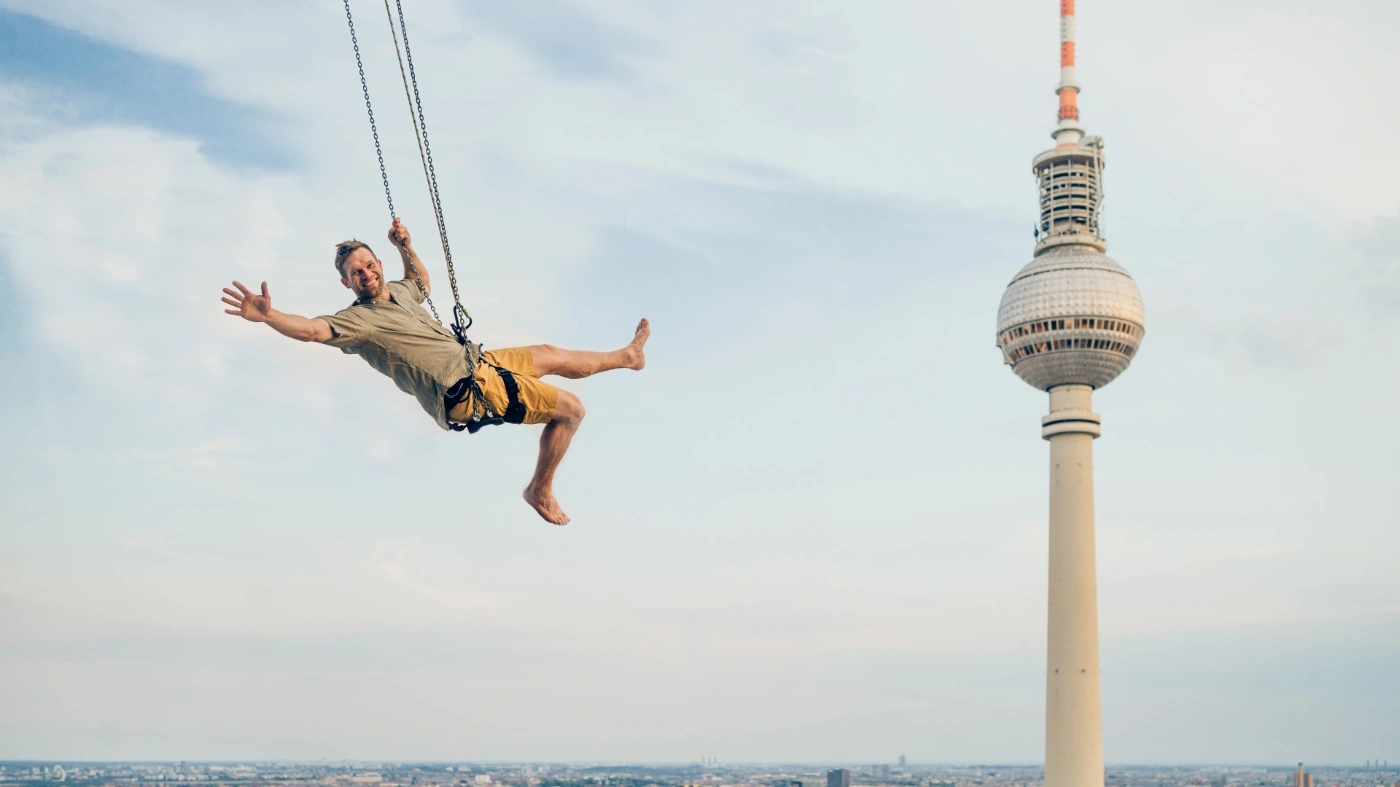 Bild einer Person die an einer langen Schaukel hoch im Himmel schwebt, im Hintergrund ist auf fast gleicher Höhe der Fernsehturm zu sehen.
