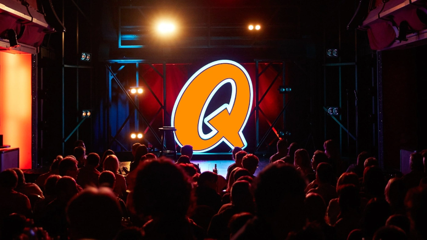 In einem vollen, abgedunkelten Saal ist auf der Bühne das große, orange erleuchtete Logo des Quatsch Comedy Clubs zu sehen.