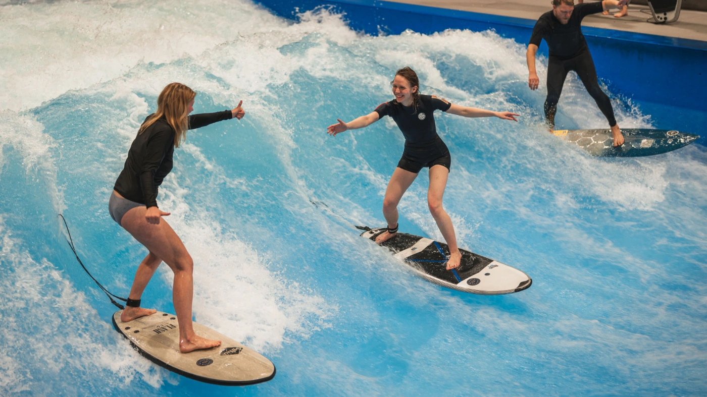 Drei Personen surfen in einer Halle auf einer künstlichen Welle.