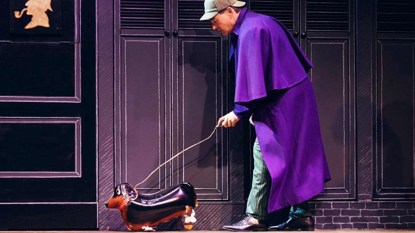 Zu sehen ist ein Schauspieler auf einer Bühne mit einem lila Mantel der Sherlock Holmes spielt. Er führt einen Hund als Ballon bei sich.