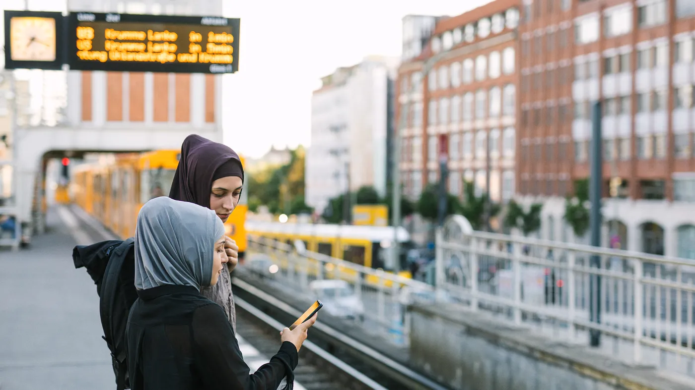 Two girls in headscarves are standing at a bus stop looking at a cell phone.