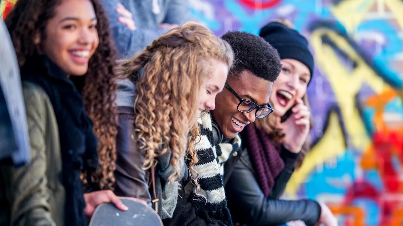 A group of young people sits laughing in front of some graffiti.