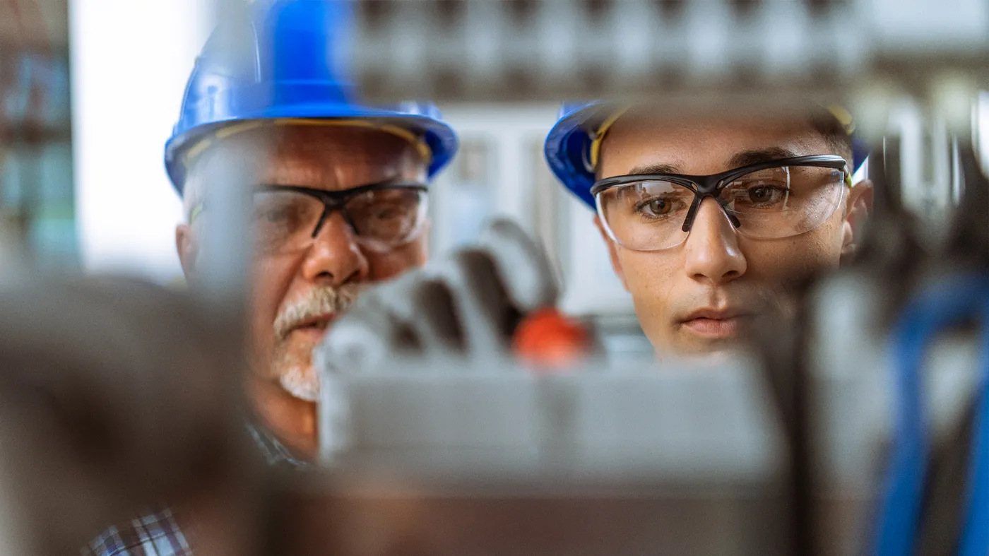 An apprentice and his foreman inspect a technical device. Both wear safety goggles and a helmet.