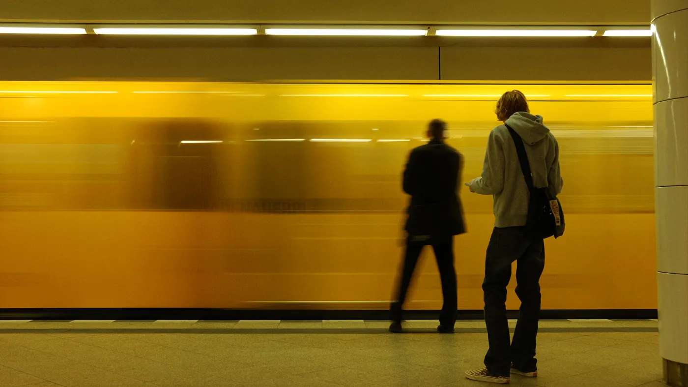 Two men are standing in a subway station. A train is just arriving and can be seen blurred in the background.