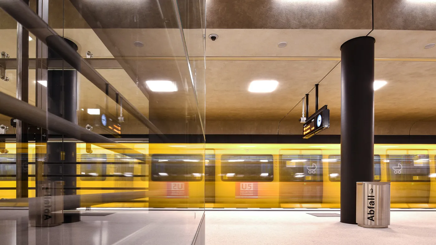 Blick über den hellen und freundlichen Bahnsteig im U-Bahnhof Unter den Linden. Im Hintergrund ist eine fahrende U-Bahn erkennbar.  