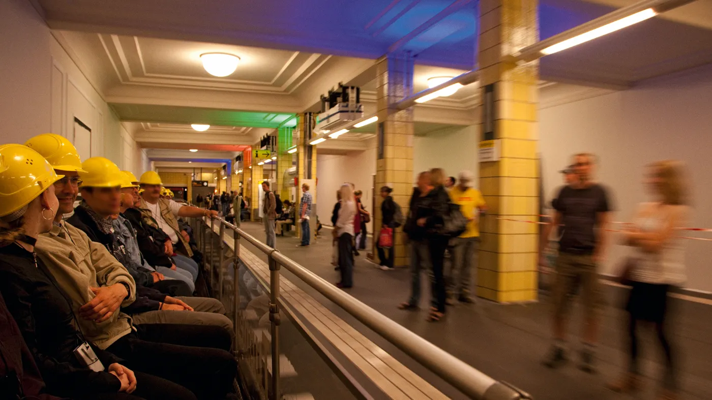 Menschen mit Schutzhelmen sitzen auf dem U-Bahn-Cabrio während es durch eine U-Bahn-Station fährt.