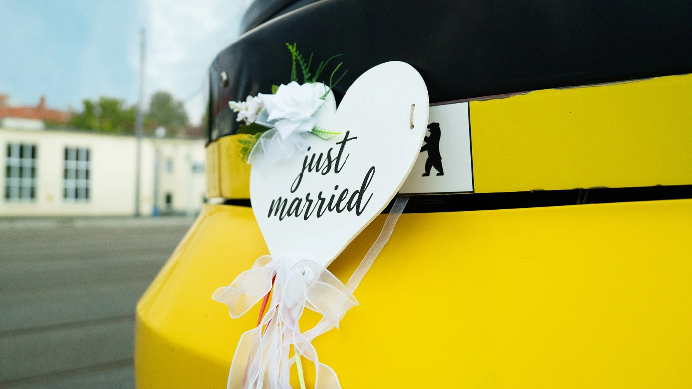 Nahaufnahme einer gelben BVG-Straßenbahn mit einem weißen, mit Blumen und Schleifen verzierten Herzschild mit der Aufschrift „just married“.