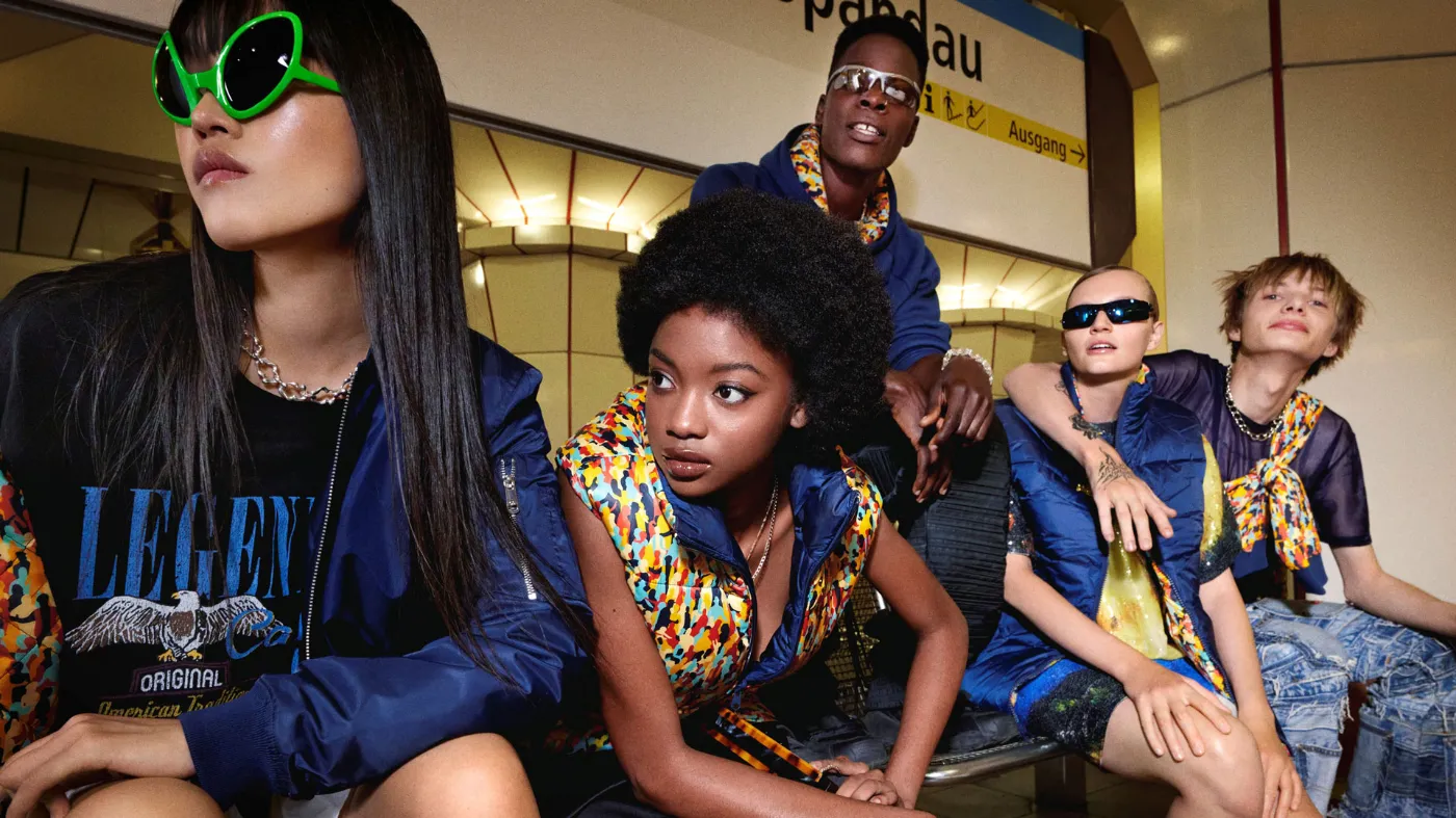 Eine Gruppe junger Menschen sitzt im U-Bahnhof auf einer Bank. Sie tragen unterschiedliche Teile der neuen BVG-Streetwear-Kollektion, die aus dem Muster der Vielfalt entstand. Einige tragen skurrile Sonnenbrillen und alle blicken in eine Richtung. Es scheint als würden sie an einem vorbeischauen. Die Szene entspricht der eines Hochglanzmotivs aus einer renommierten Modezeitung.