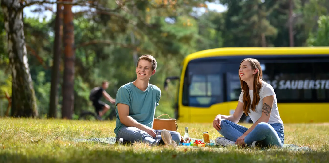 En junges Paar picknickt auf einer Wiese. Im Hintergrund fährt ein Bus der BVG vorbei. 